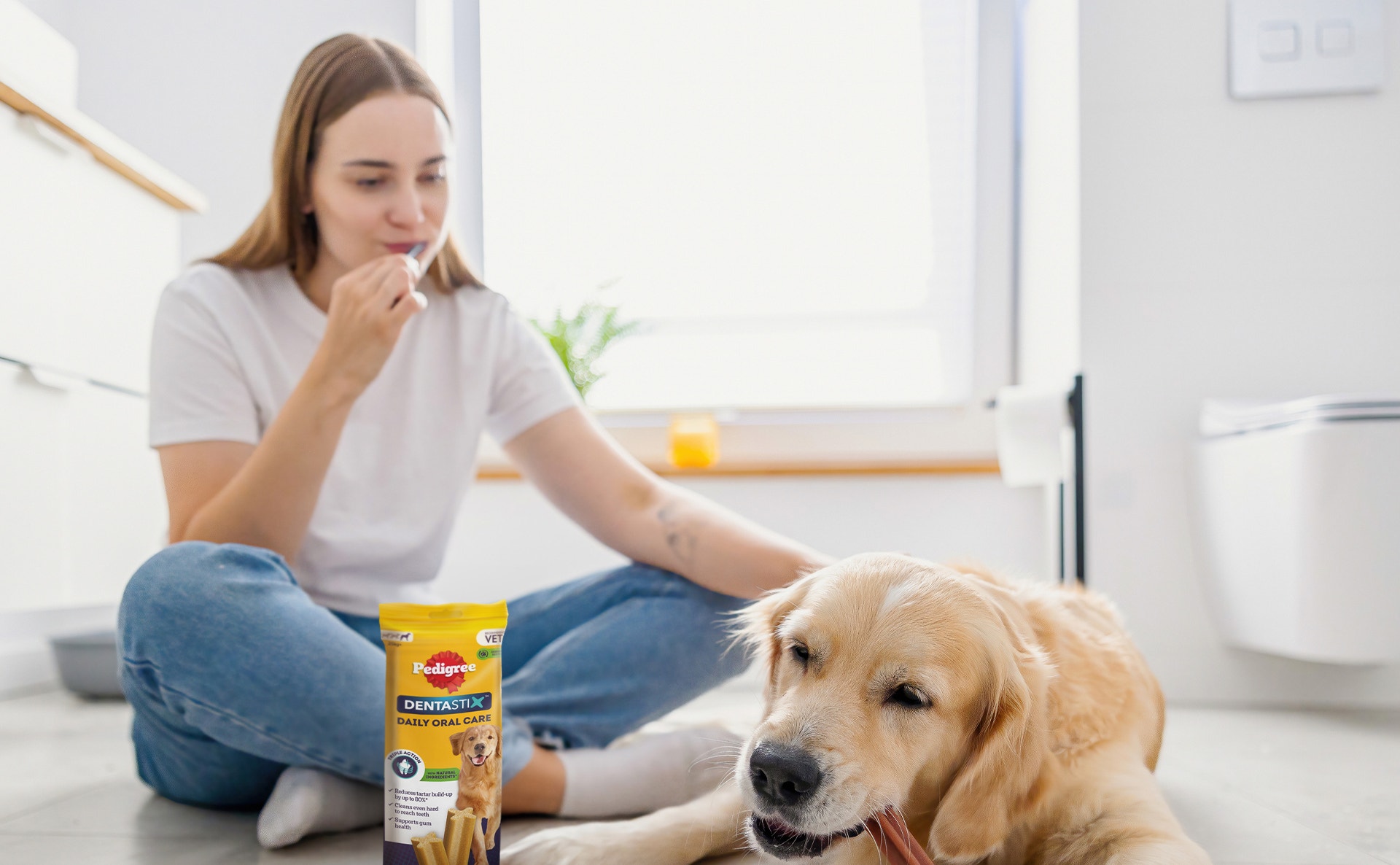 Une jeune femme est assise sur le sol avec un golden retriever qui mâche un bâtonnet Pedigree Dentastix. Un paquet de Dentastix est posé à côté d'eux.